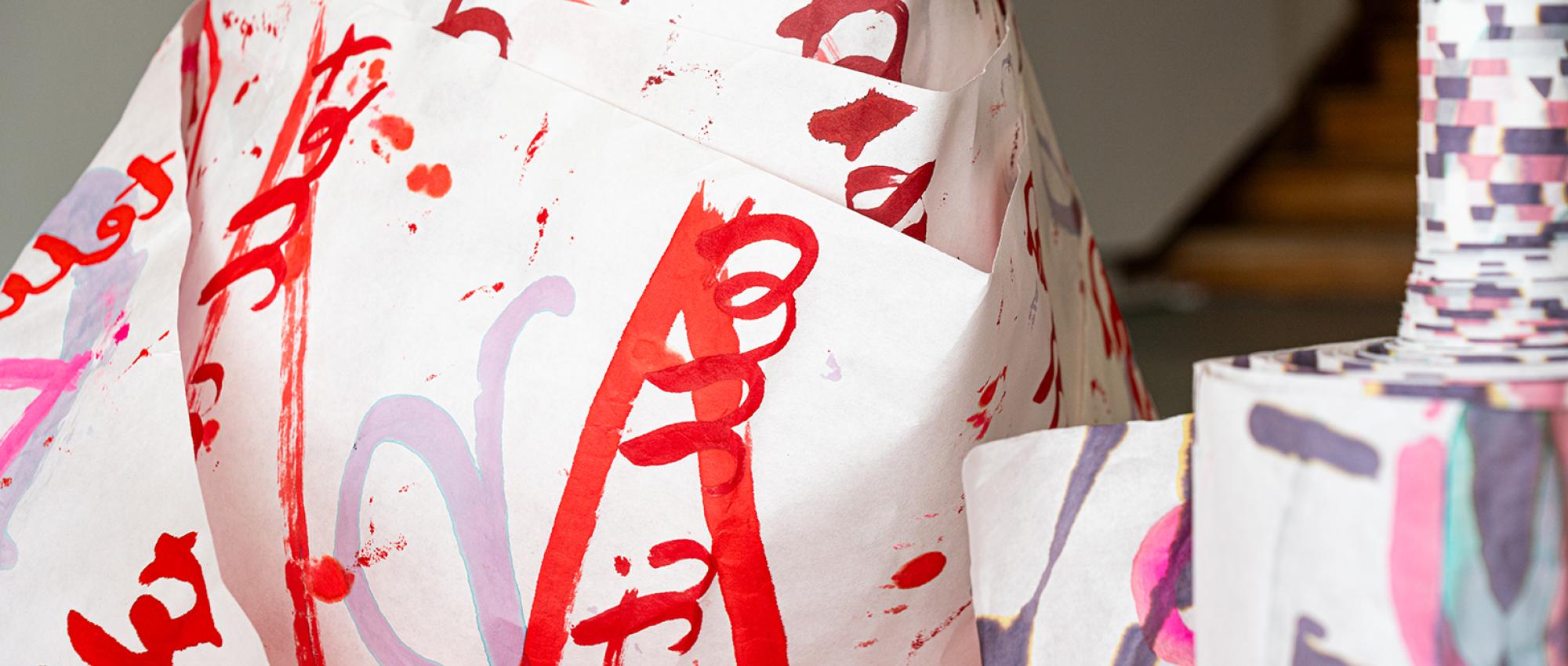Close-up shot of two paper scrolls standing upright in a gallery. The paper is painted with text and letter-like forms. If you perform some mental gymnastics you can maybe make out some words painted backwards.